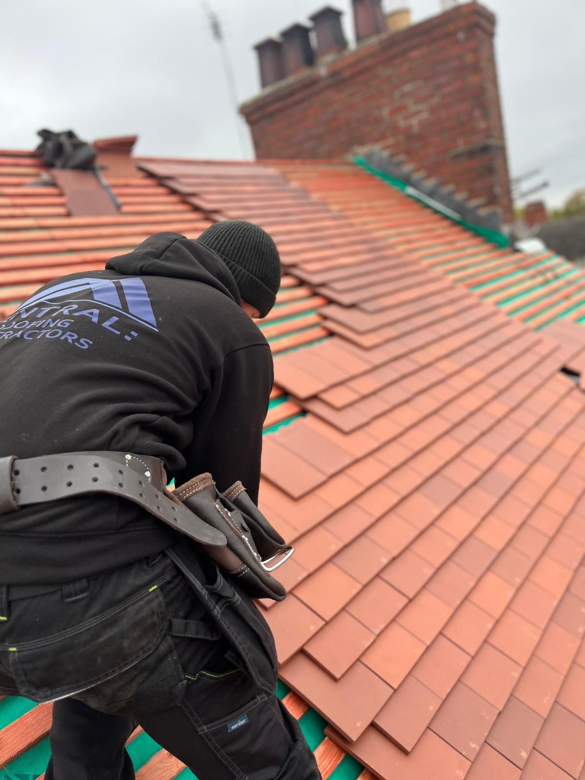 Roofer laying red tiles on a slanted roof next to a brick chimney under an overcast sky.