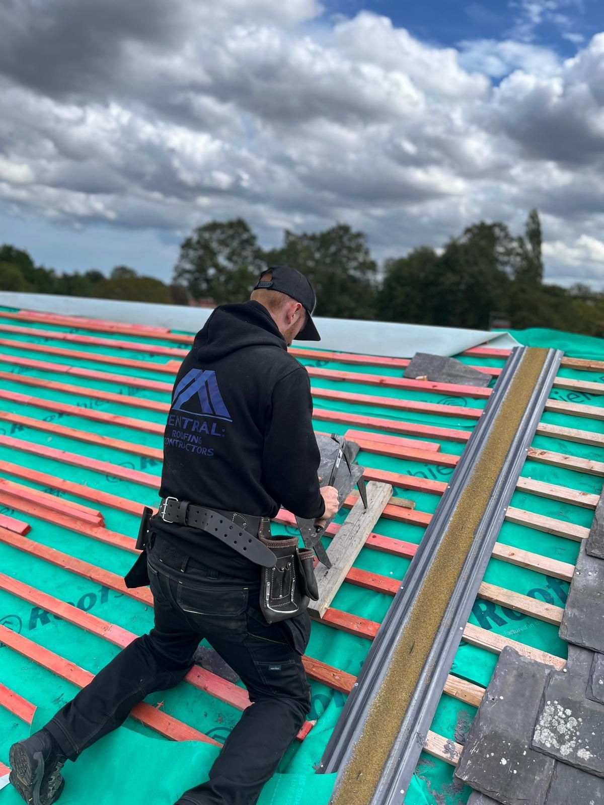 Roofer on a roof cutting tiles. Wearing a black hoodie, using a saw. Cloudy sky in the background.