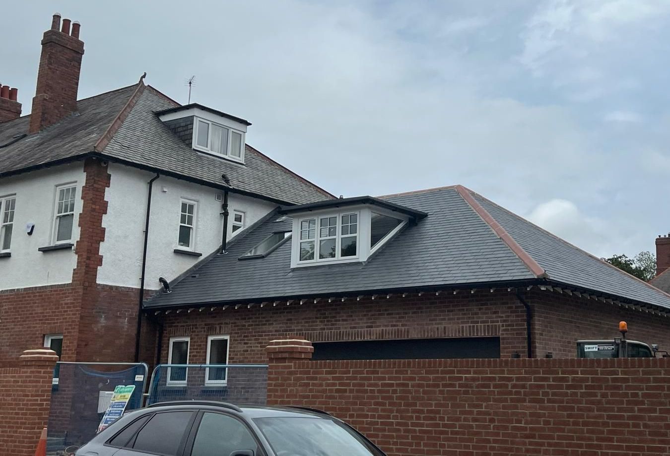 Building with slate roof, red brick, and white trim. Grey car and brick wall in foreground. Overcast sky.