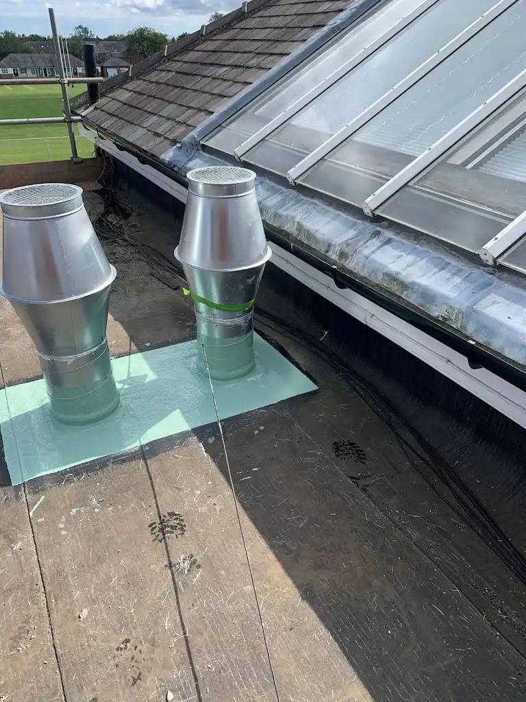 Two silver roof vents on a flat roof with a skylight. Green insulation pads and black roofing material.