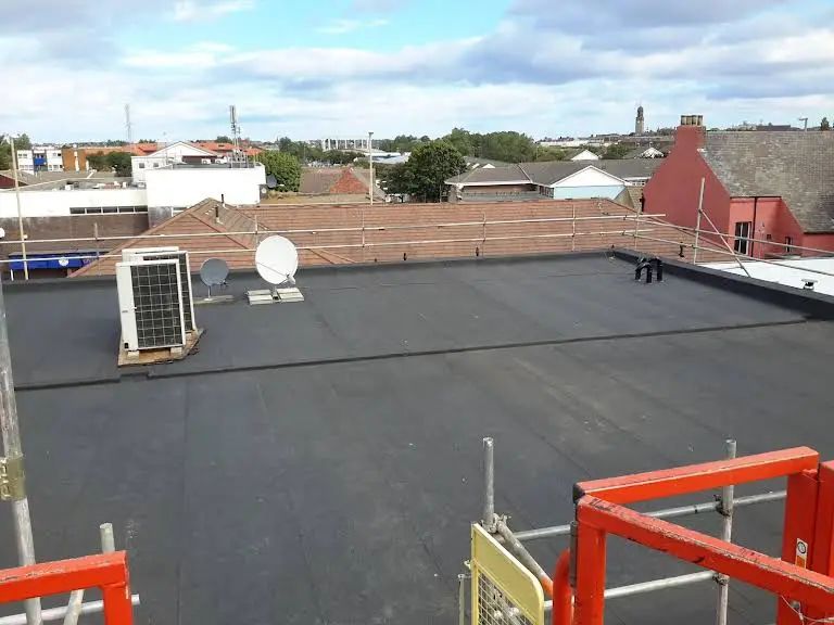 Rooftop with black surface, air conditioning unit, satellite dish, and surrounding buildings under a cloudy sky.