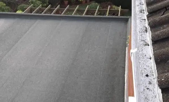 Flat, dark roof surface next to a gutter filled with debris and a tiled roof.