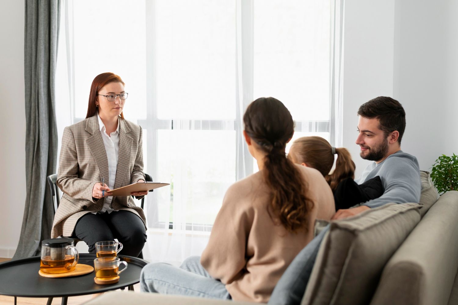 Therapist speaking with three clients in a bright living room, seated on a sofa with tea on the table.