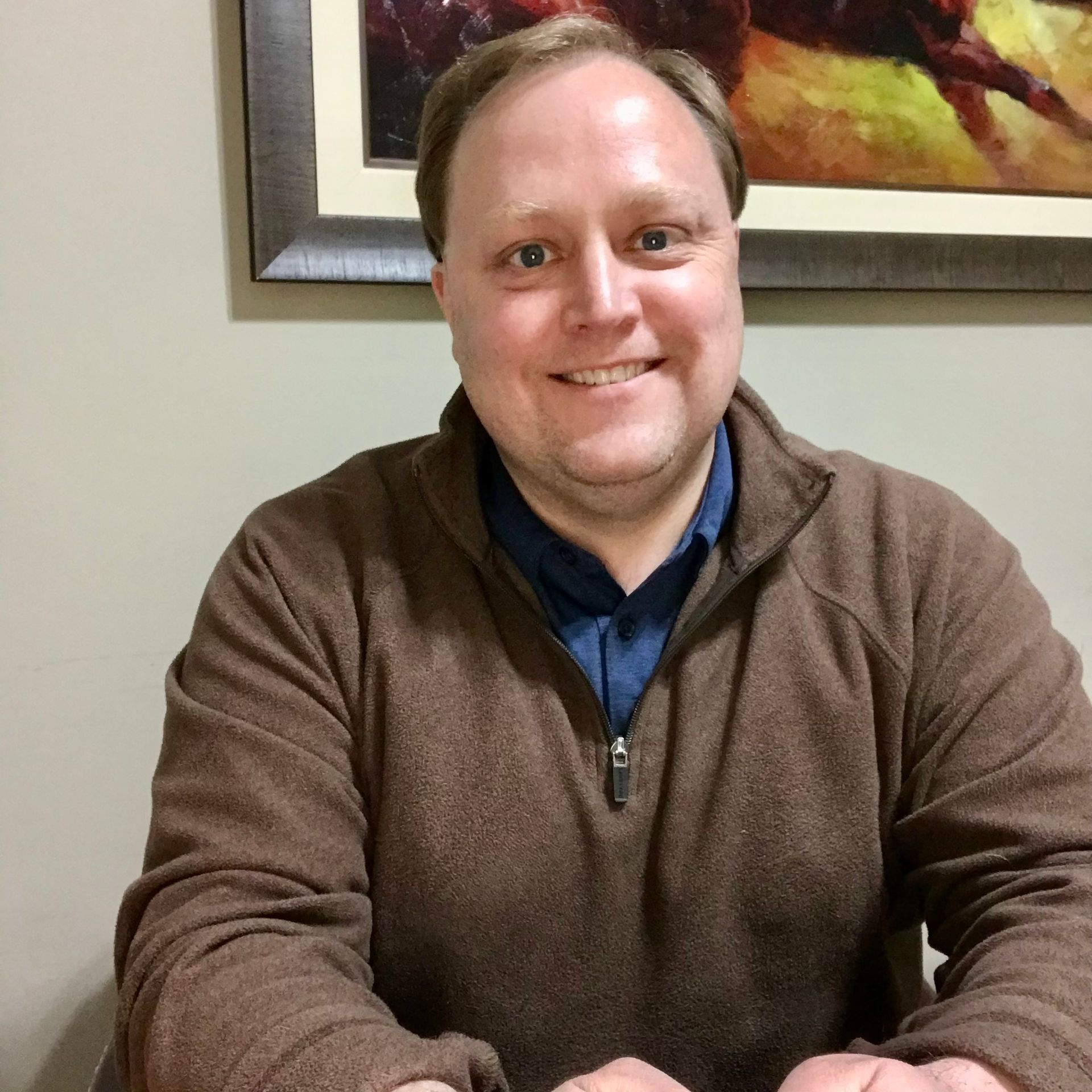 Smiling person in a brown sweater seated indoors in front of a framed wall picture.
