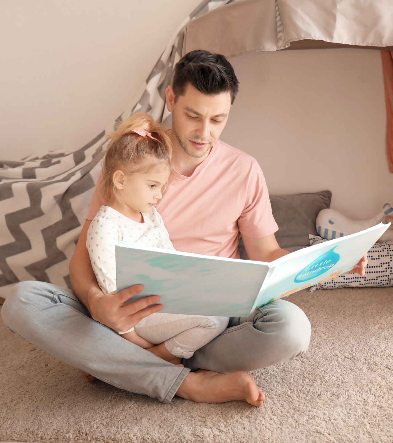 Adult and child sitting on a carpet reading a book inside a cozy tent, with soft cushions nearby