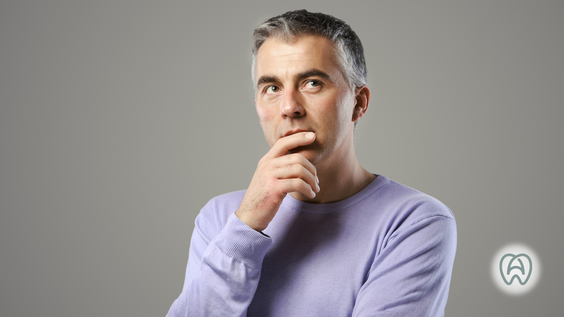 Man in a lavender sweater touching his chin, looking thoughtfully at the camera against a gray background
