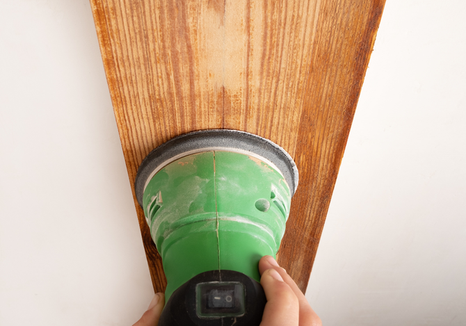 A person is sanding a piece of wood with a green sander.