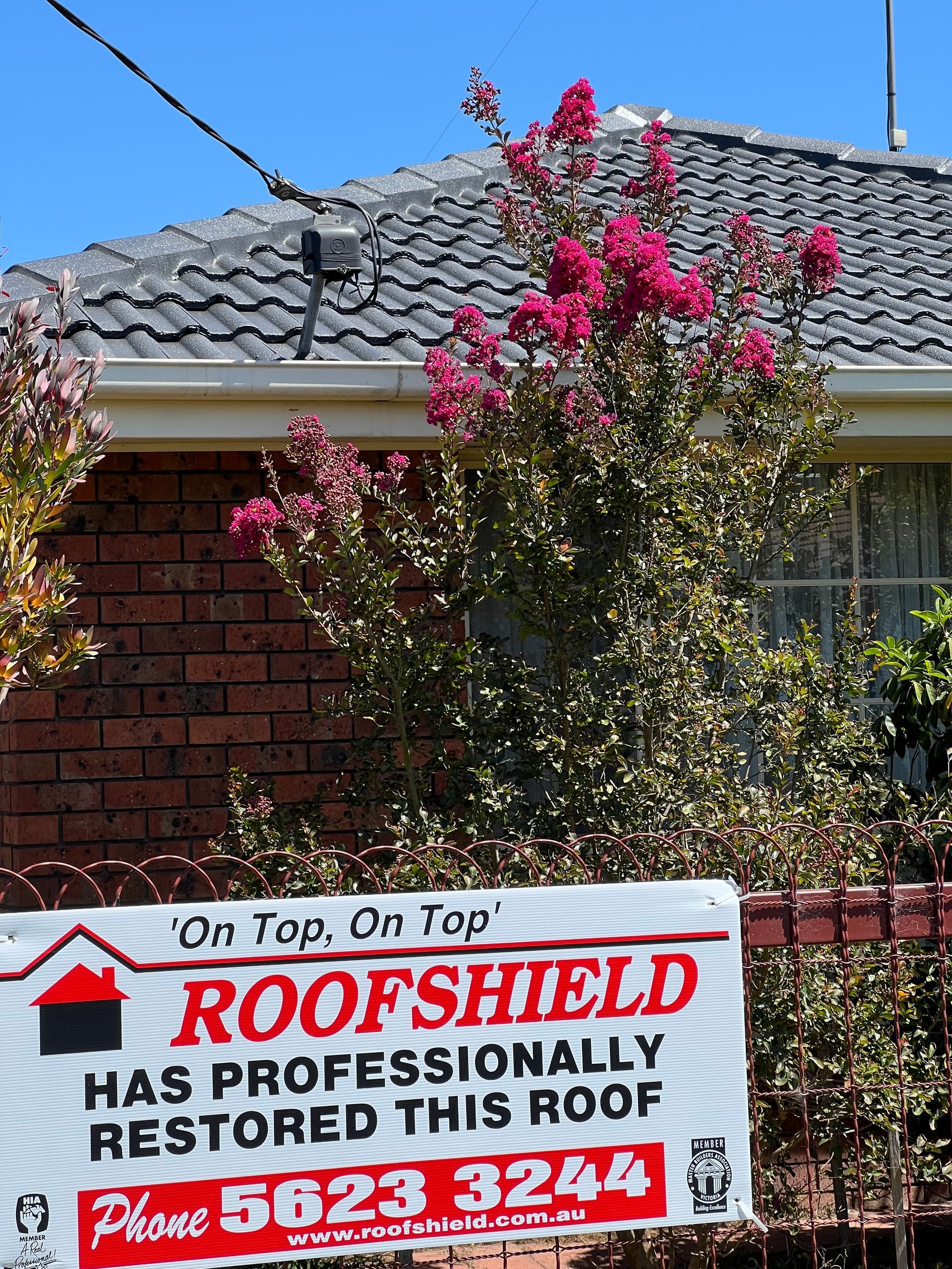 Roof restoration sign in front of a brick house with a blooming pink tree.