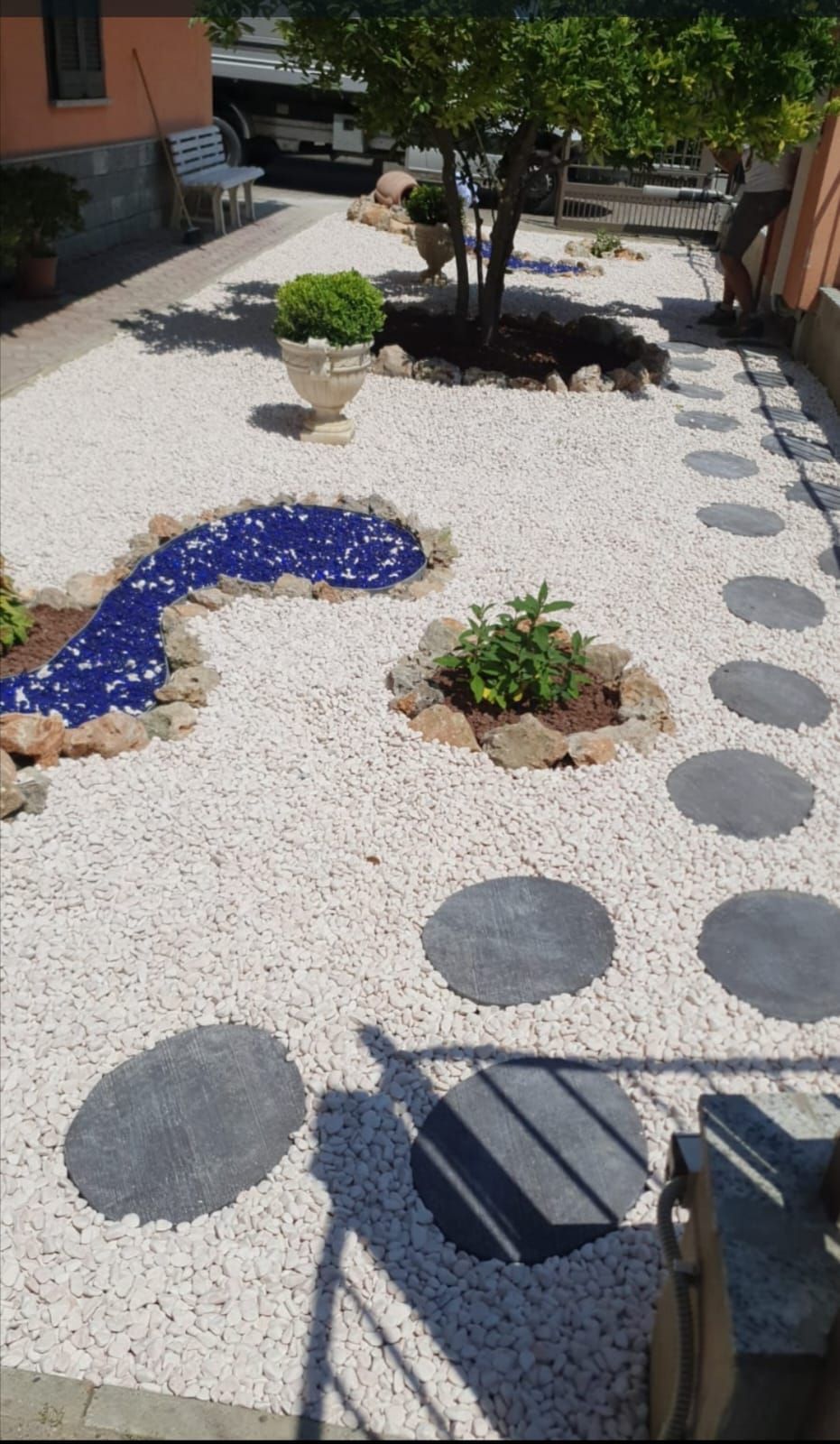 Sentiero da giardino in ghiaia bianca con pietre rotonde da calpestare, piante in vaso e un piccolo laghetto decorativo blu.