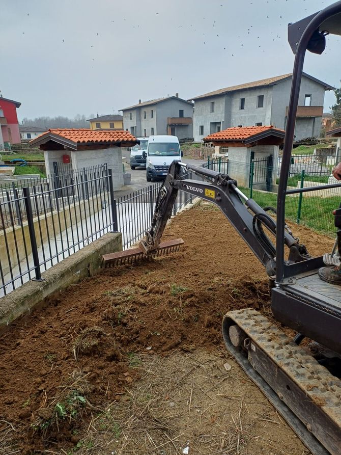 Un escavatore sta scavando una trincea accanto a una strada residenziale recintata, con cumuli di terra lungo il percorso.