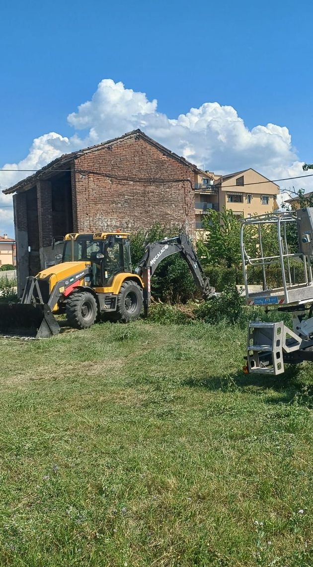 Una terna gialla in un cortile erboso accanto a un vecchio edificio in mattoni, sotto un cielo azzurro con nuvole.