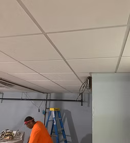 Worker on a ladder in a room with a drop ceiling, installing or repairing ceiling tiles and wiring.