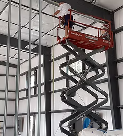 Worker on a red scissor lift installing a metal frame inside a steel building.