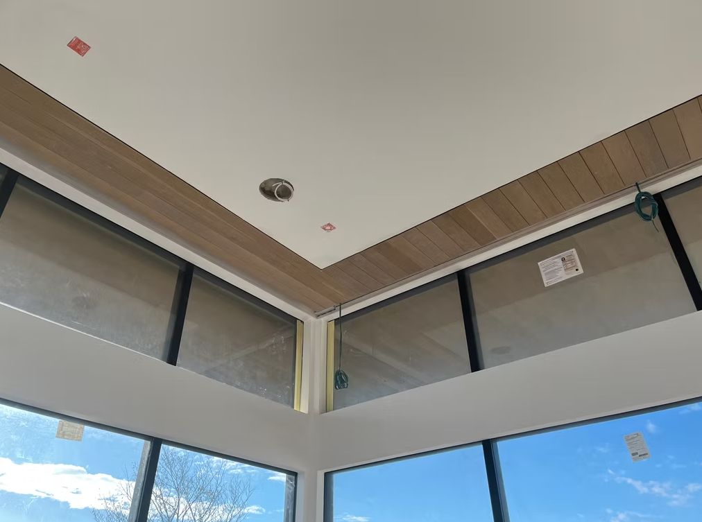 Corner of a modern room with high windows, wood trim, white ceiling, and blue sky outside.