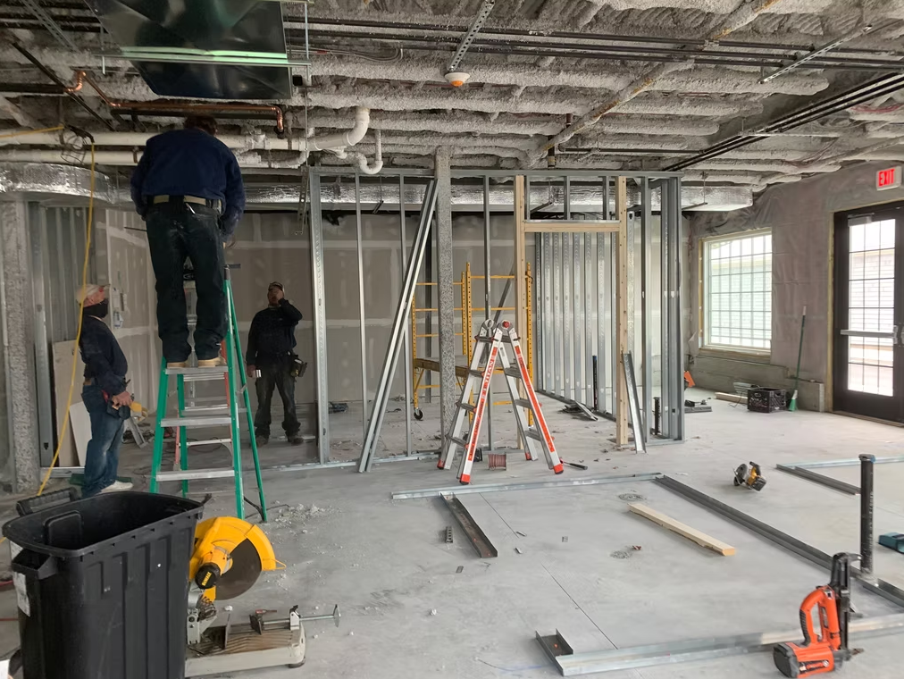 Construction workers in unfinished room with ladders, tools, and exposed framing and ceiling fixtures
