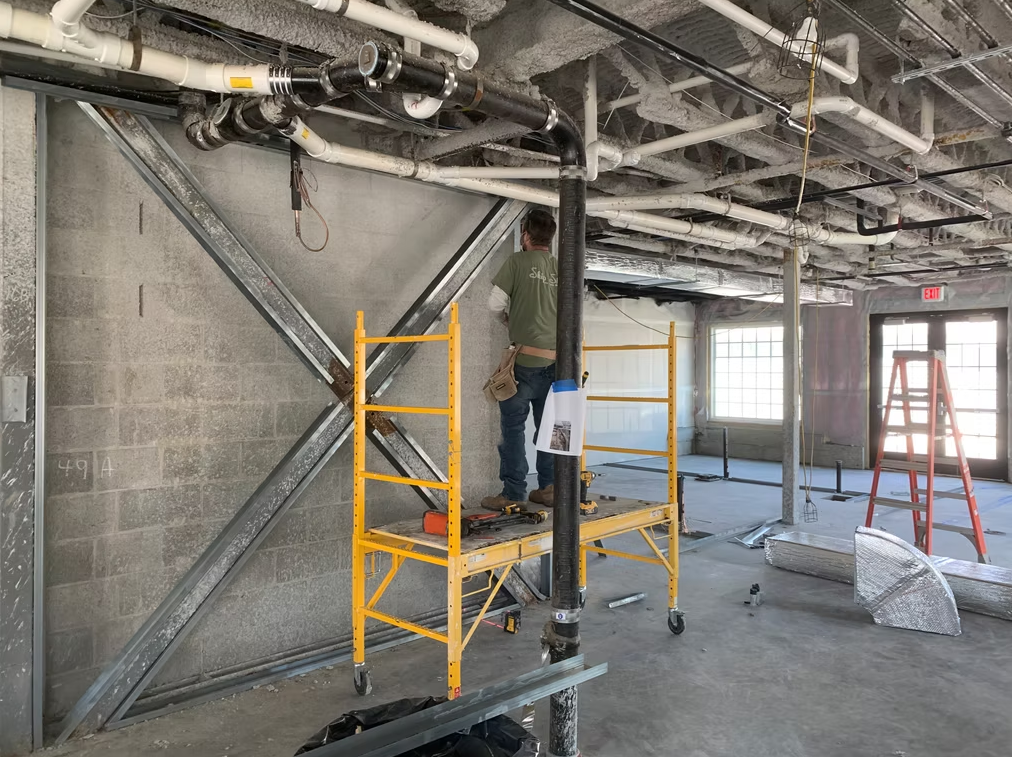 Worker on yellow scaffolding in a concrete room with exposed pipes and unfinished ceiling.