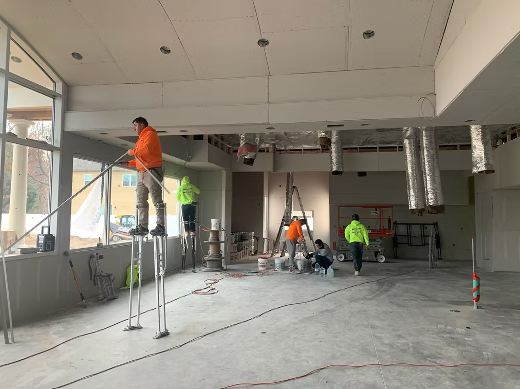 Construction workers in a bright unfinished room, one on a ladder near windows and another on scaffolding.