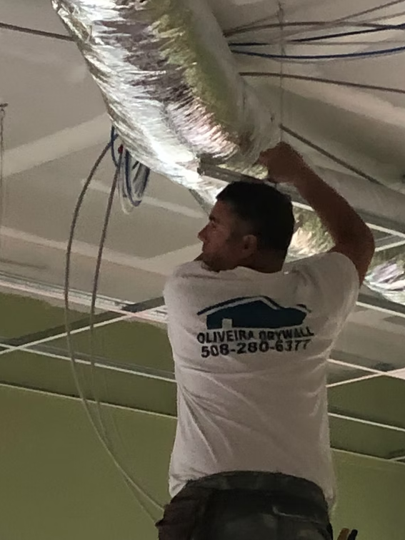 Worker installing ceiling ductwork in a room with exposed wires and a green wall.