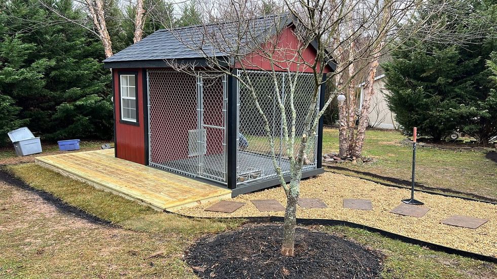 Small red garden shed with wire-mesh enclosure on a paved path in a yard with trees and grass