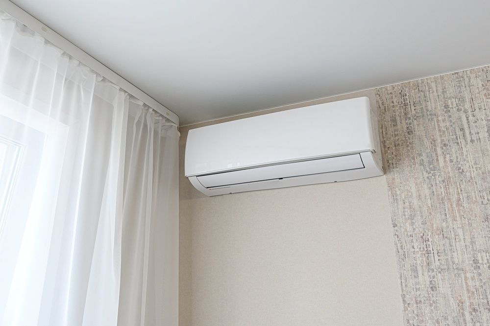 White wall-mounted air conditioner above a window with sheer curtains in a bright room — GSA Group In Garbutt, QLD