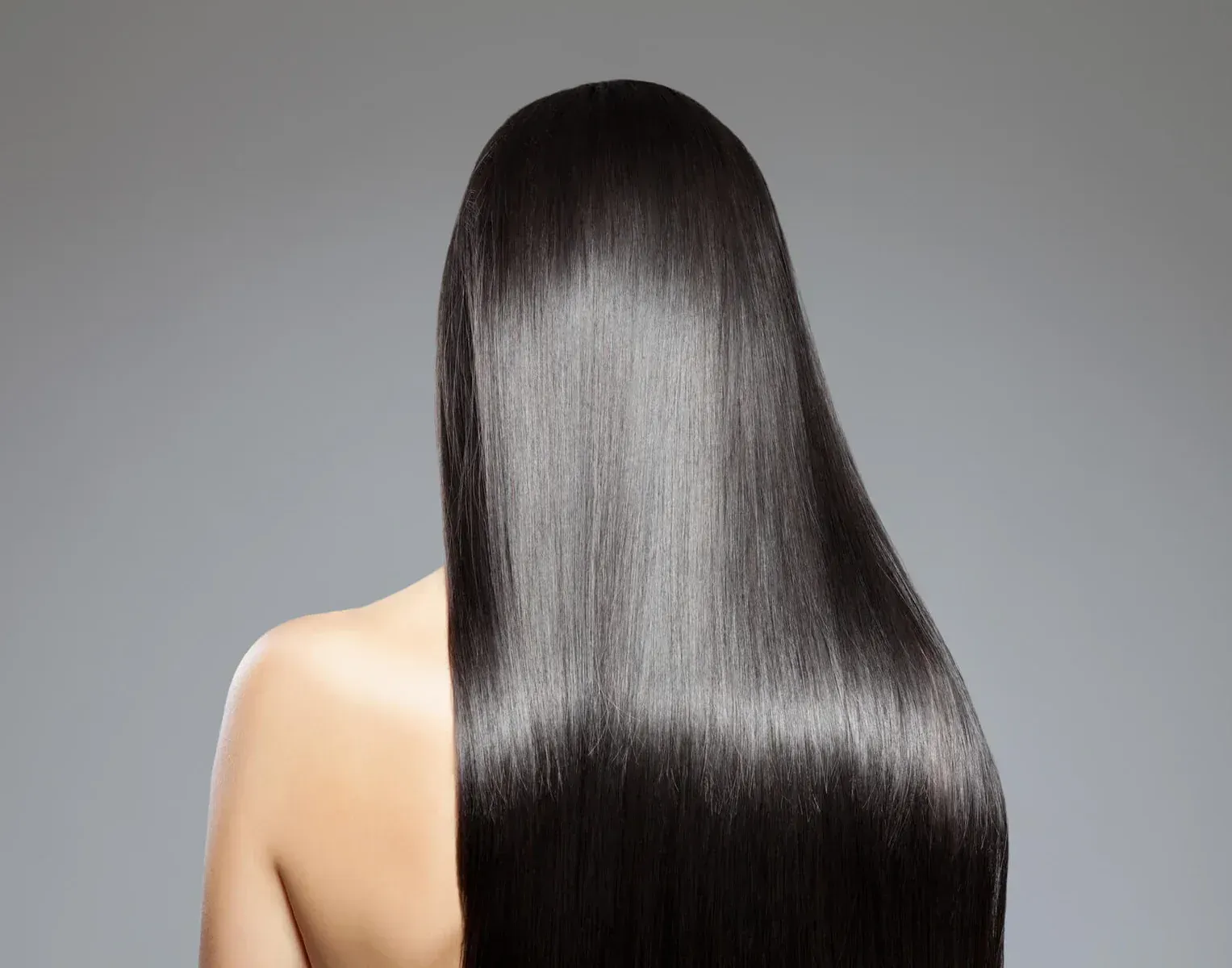 Mujer con cabello largo, brillante, negro y liso, visto de espaldas, sobre un fondo gris.