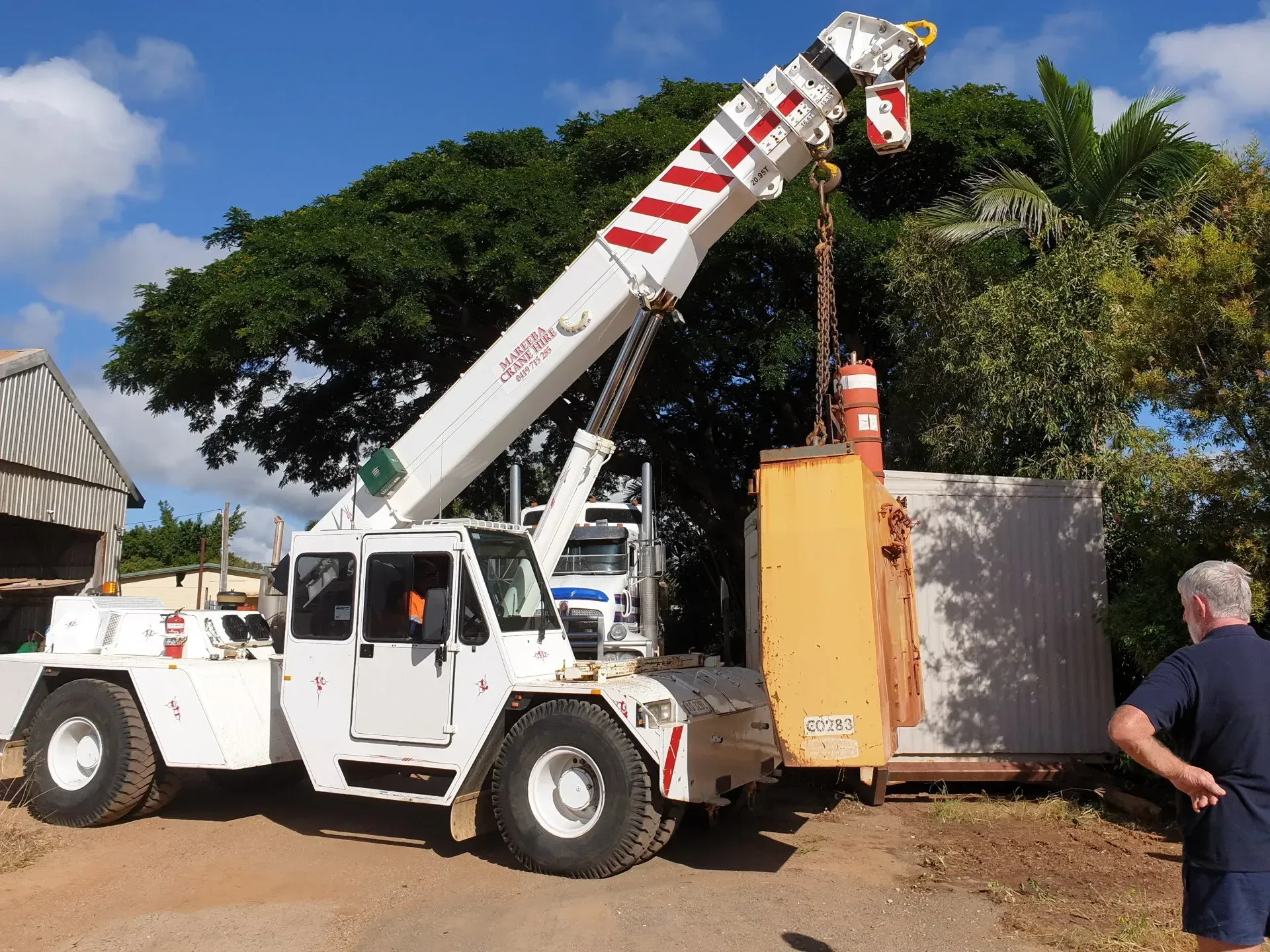 A White Crane Lifts a Yellow Industrial Container Next to a Metal Structure — Mareeba Crane Hire In Mareeba, QLD