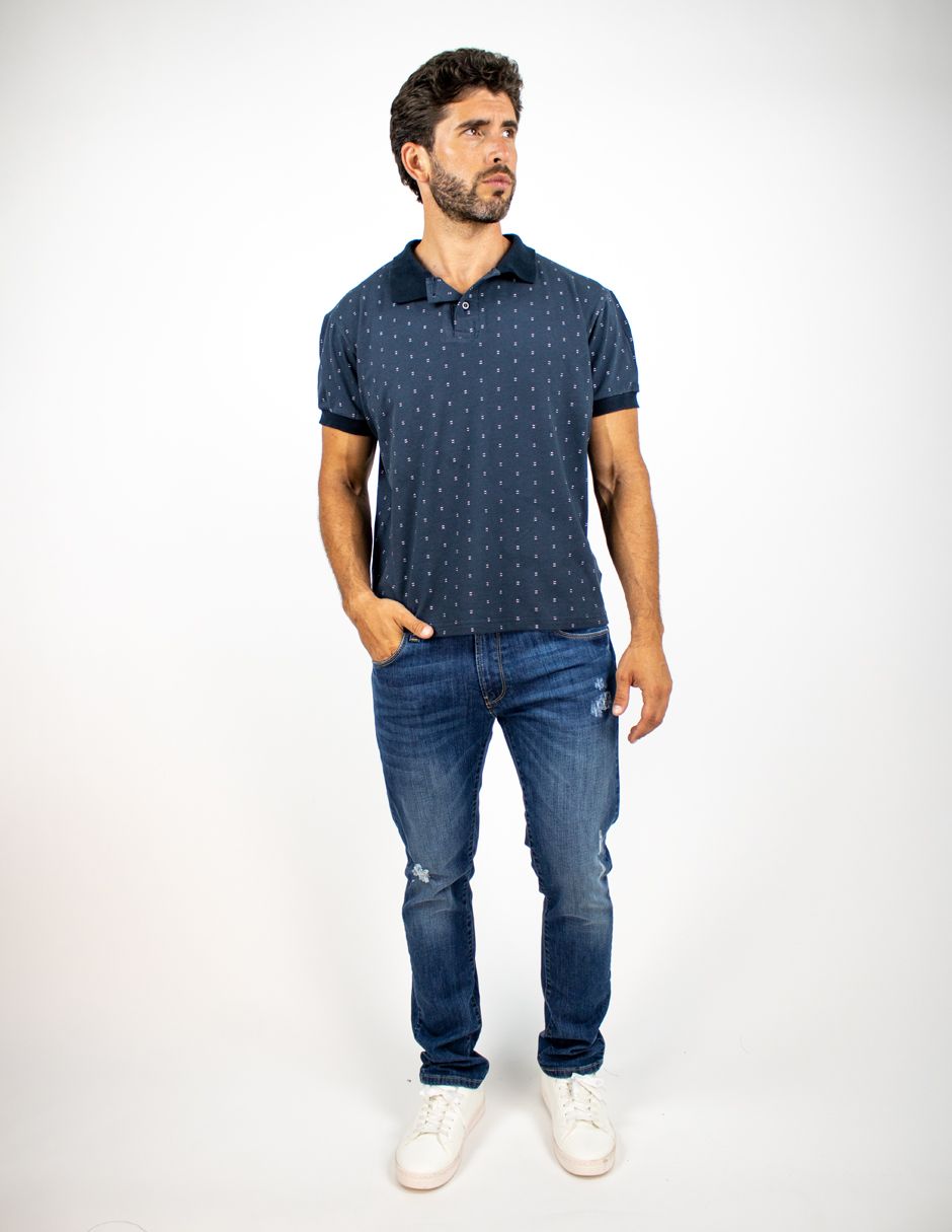 Hombre de pie con una camisa polo azul estampada, vaqueros y zapatillas blancas sobre un fondo blanco.