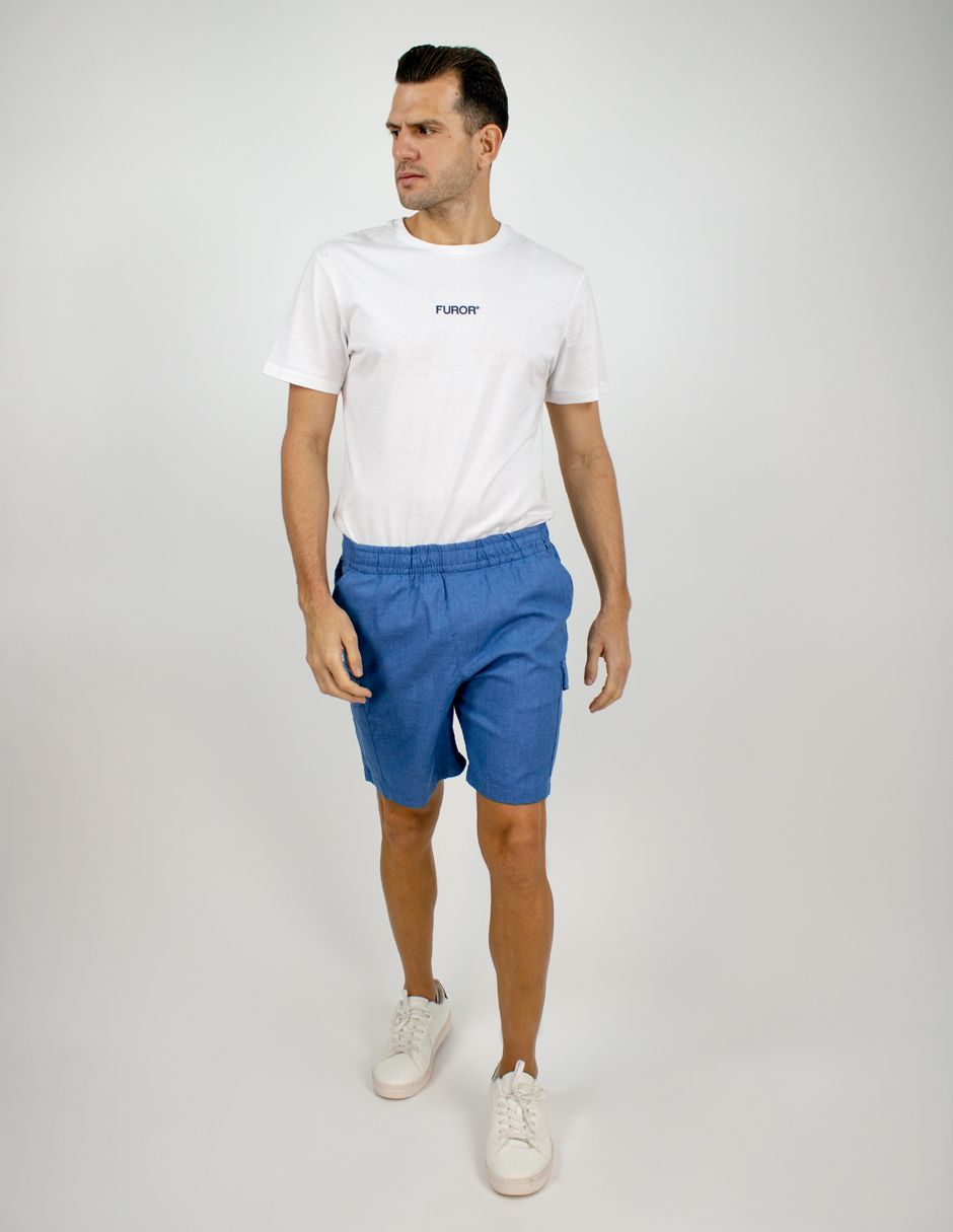 Hombre de pie con camiseta blanca, pantalones cortos azules y zapatillas blancas sobre un fondo liso.