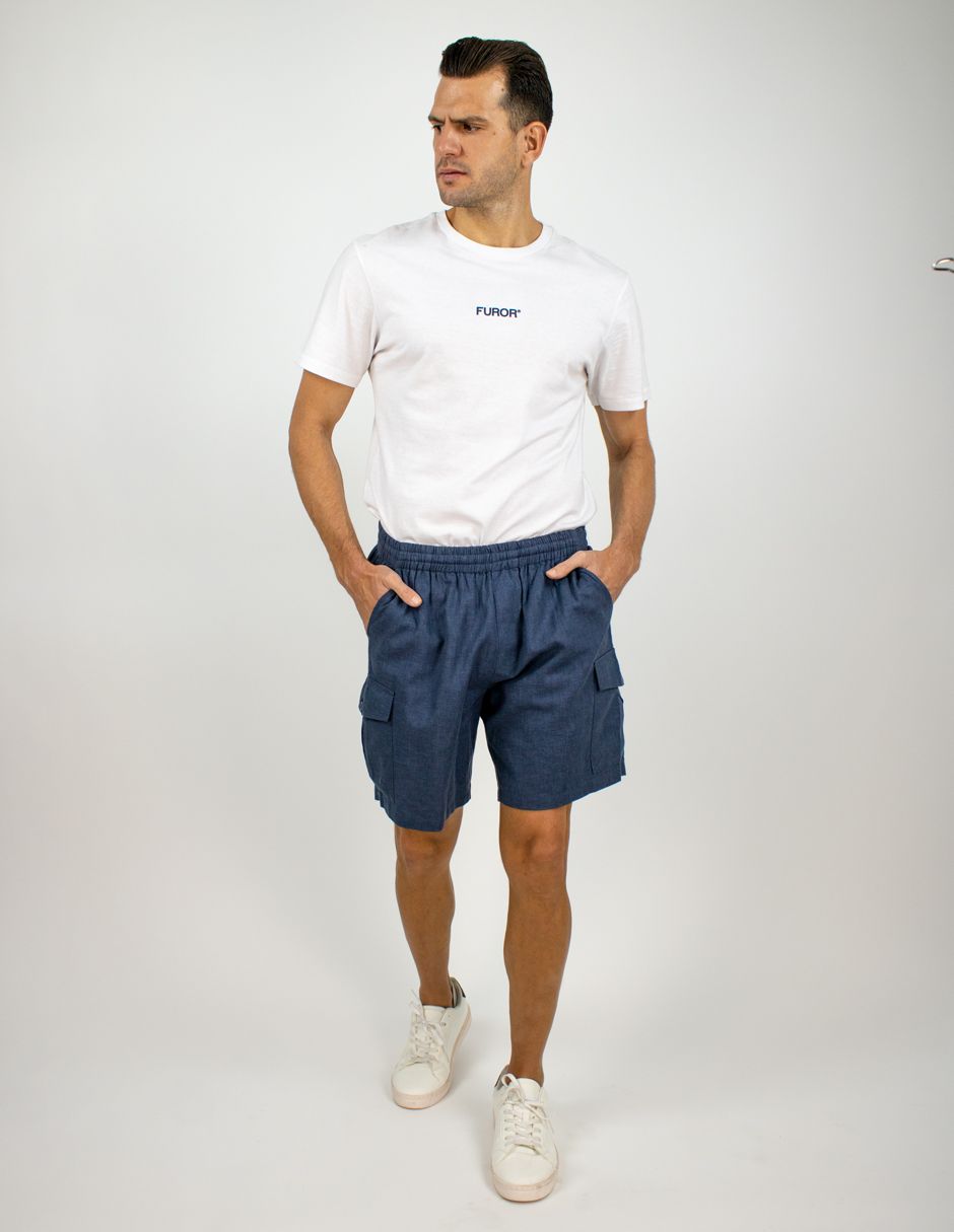 Hombre de pie con camiseta blanca, pantalones cortos azul marino y zapatillas blancas sobre un fondo blanco liso.