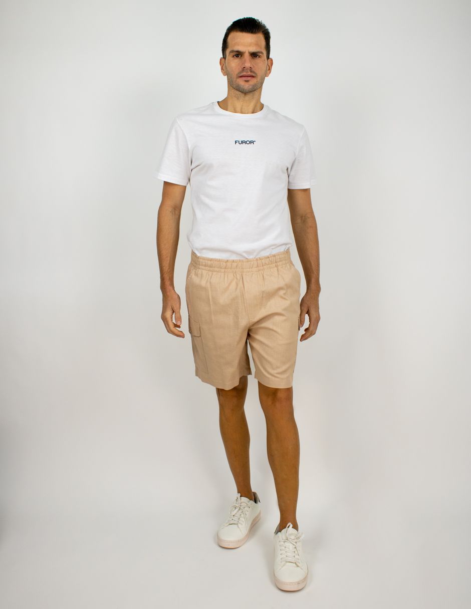 Hombre de pie sobre un fondo gris liso, vistiendo una camiseta blanca, pantalones cortos beige y zapatillas blancas.