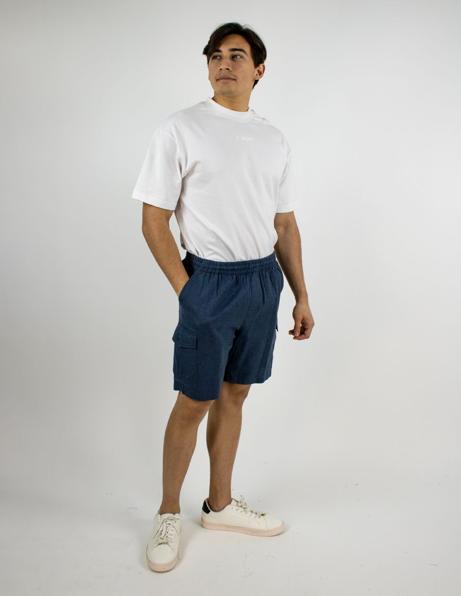 Persona vestida con camisa blanca, pantalones cortos azul marino y zapatillas blancas, de pie sobre un fondo blanco liso.