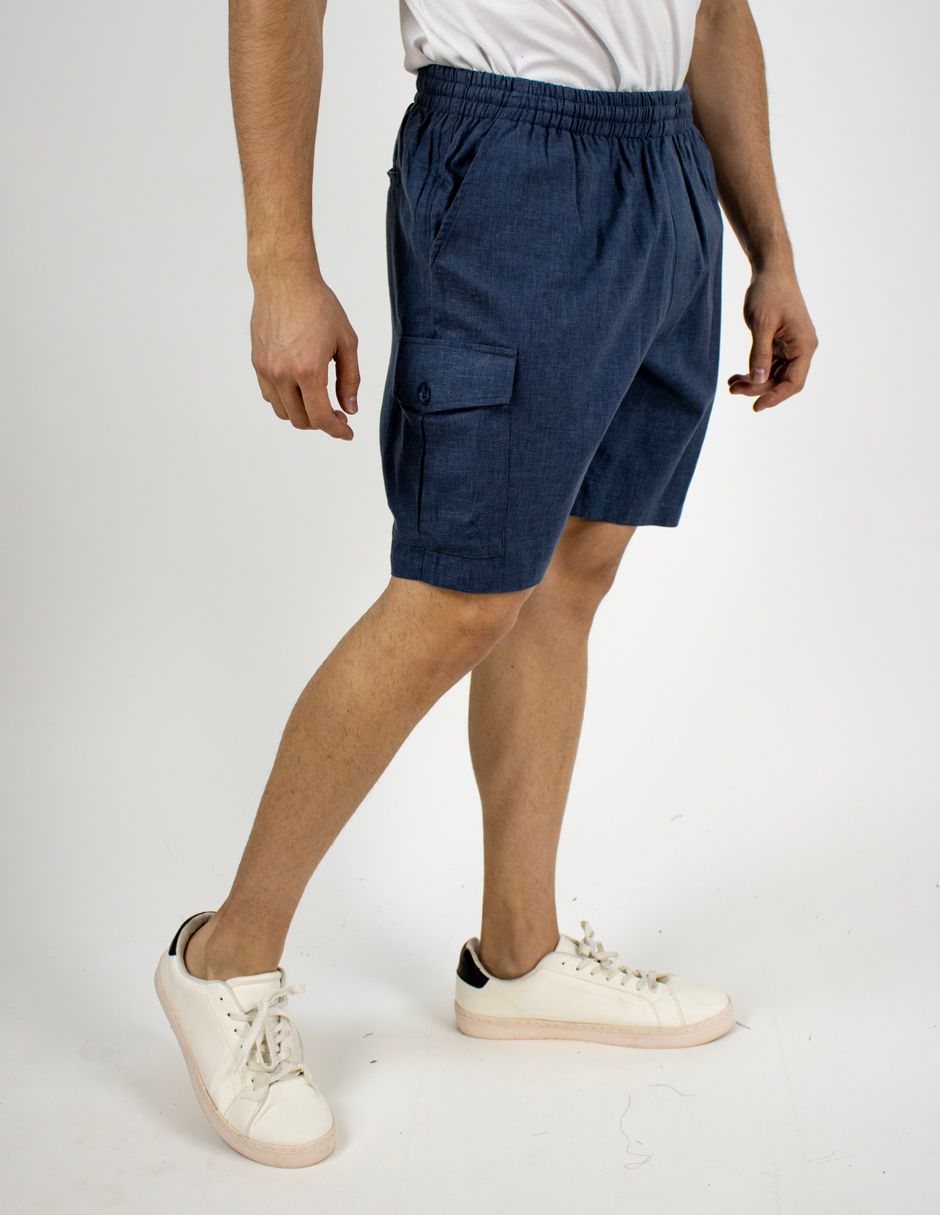 Hombre con pantalones cortos cargo azul marino, camiseta blanca y zapatillas blancas, de pie sobre un fondo liso.