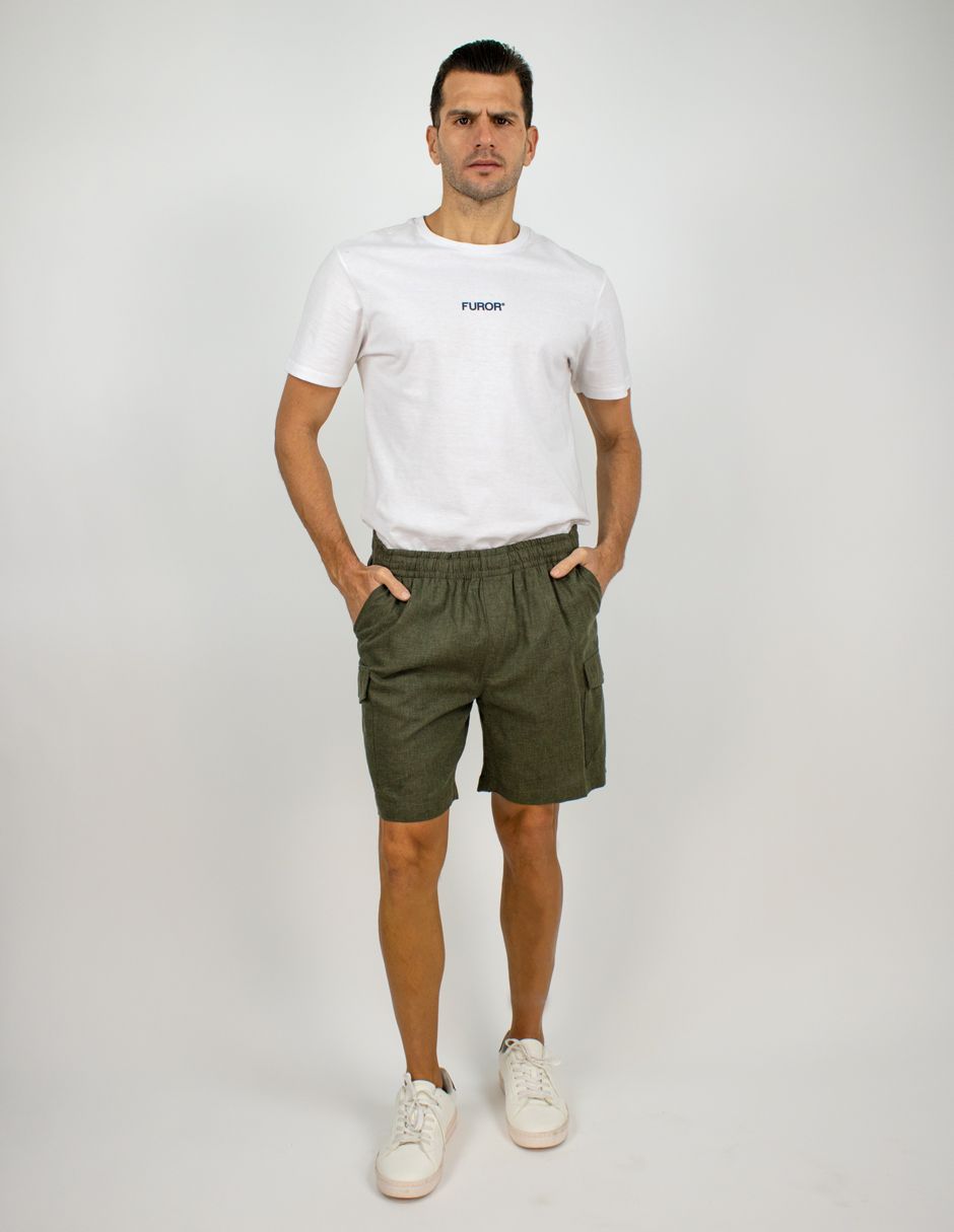 Hombre de pie sobre un fondo blanco, vistiendo una camiseta blanca, pantalones cortos color oliva y zapatillas blancas.