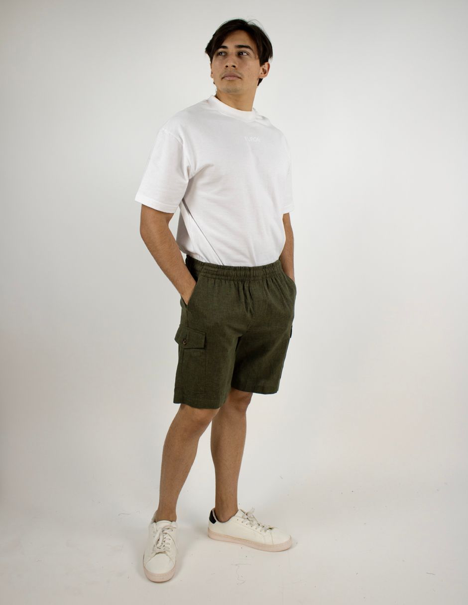 Persona de pie con una camiseta blanca, pantalones cortos color oliva y zapatillas blancas sobre un fondo blanco liso.