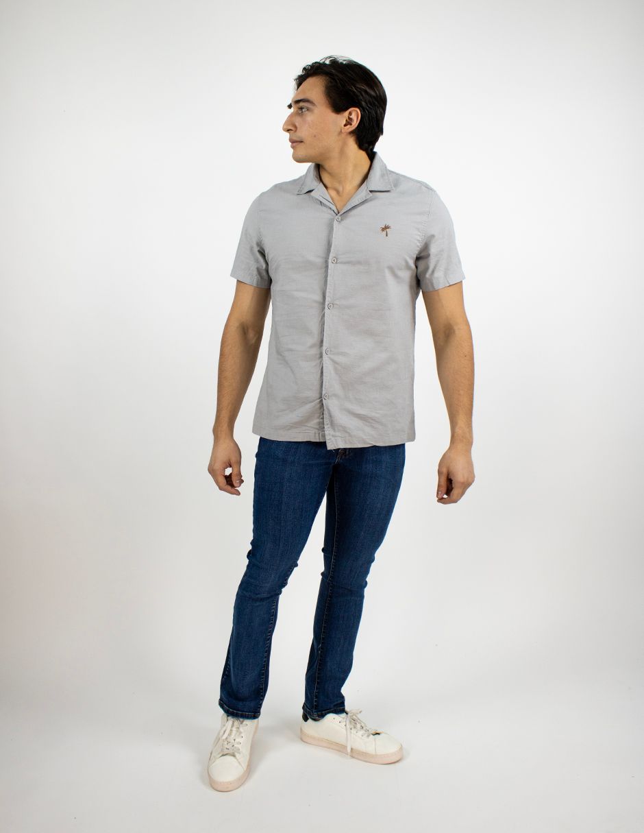 Persona de pie sobre un fondo blanco, vestida con una camisa gris de manga corta, vaqueros azules y zapatillas blancas, mirando hacia la izquierda.