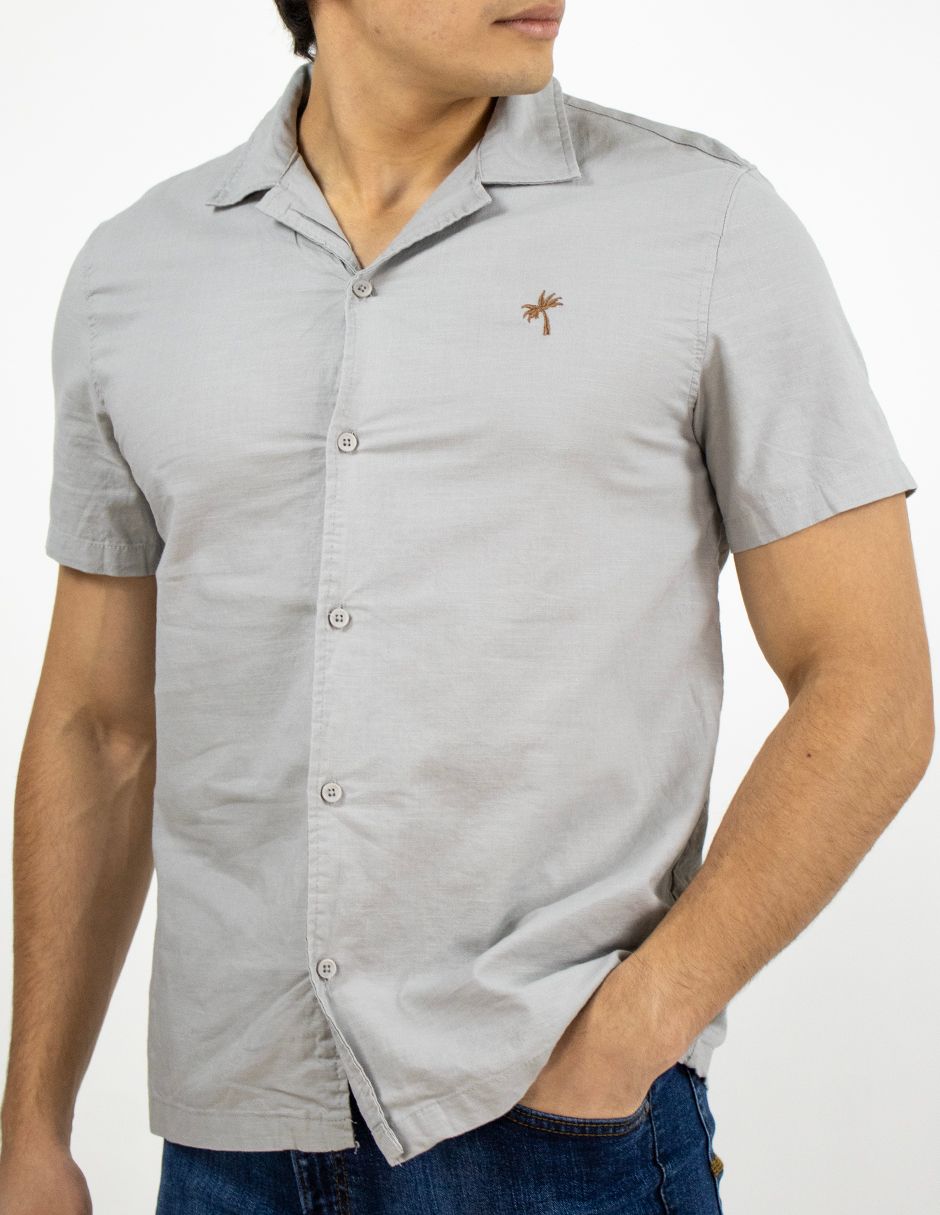 Hombre con camisa gris claro de manga corta abotonada y vaqueros, de pie con una mano en el bolsillo.