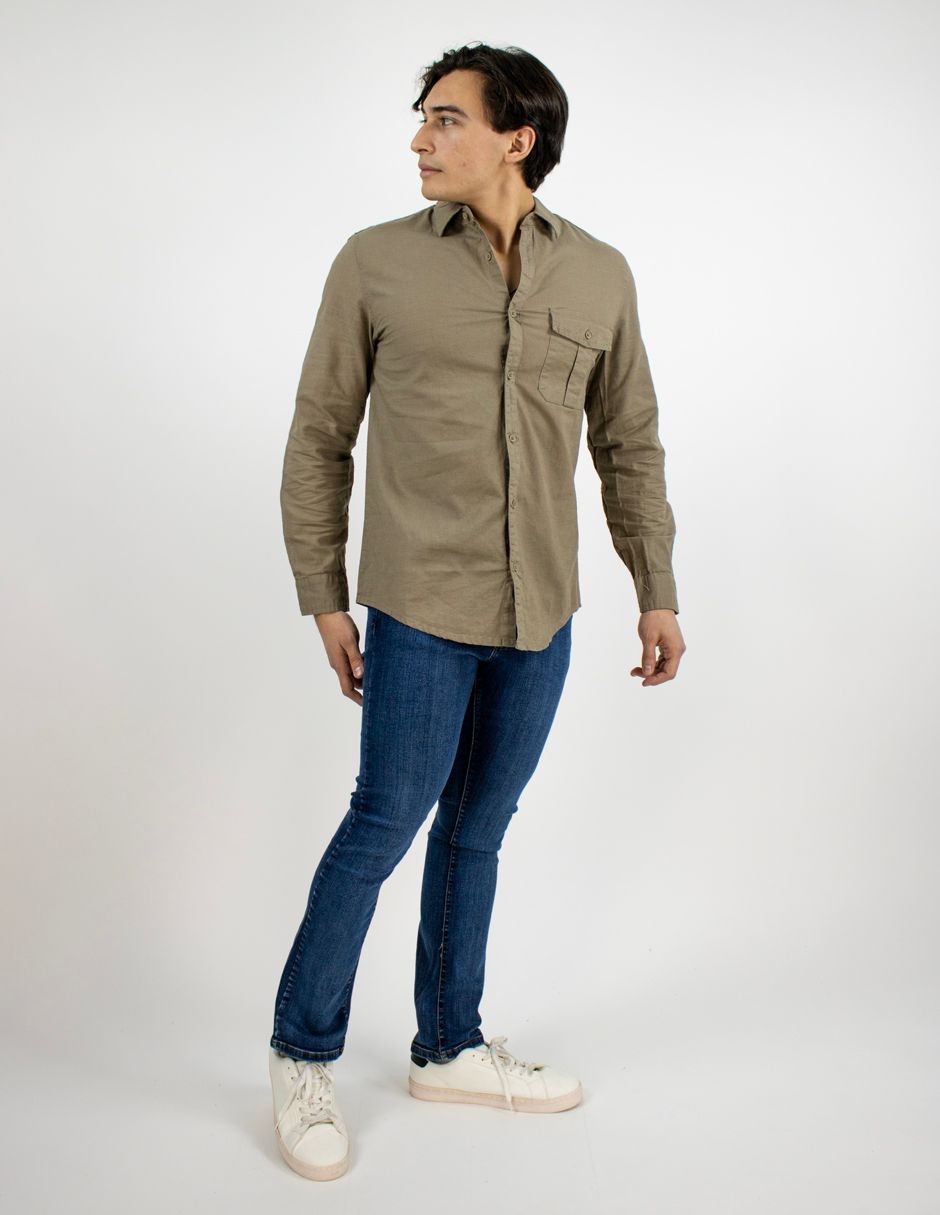 Una persona que viste una camisa color canela, vaqueros azules y zapatillas blancas posa sobre un fondo blanco.