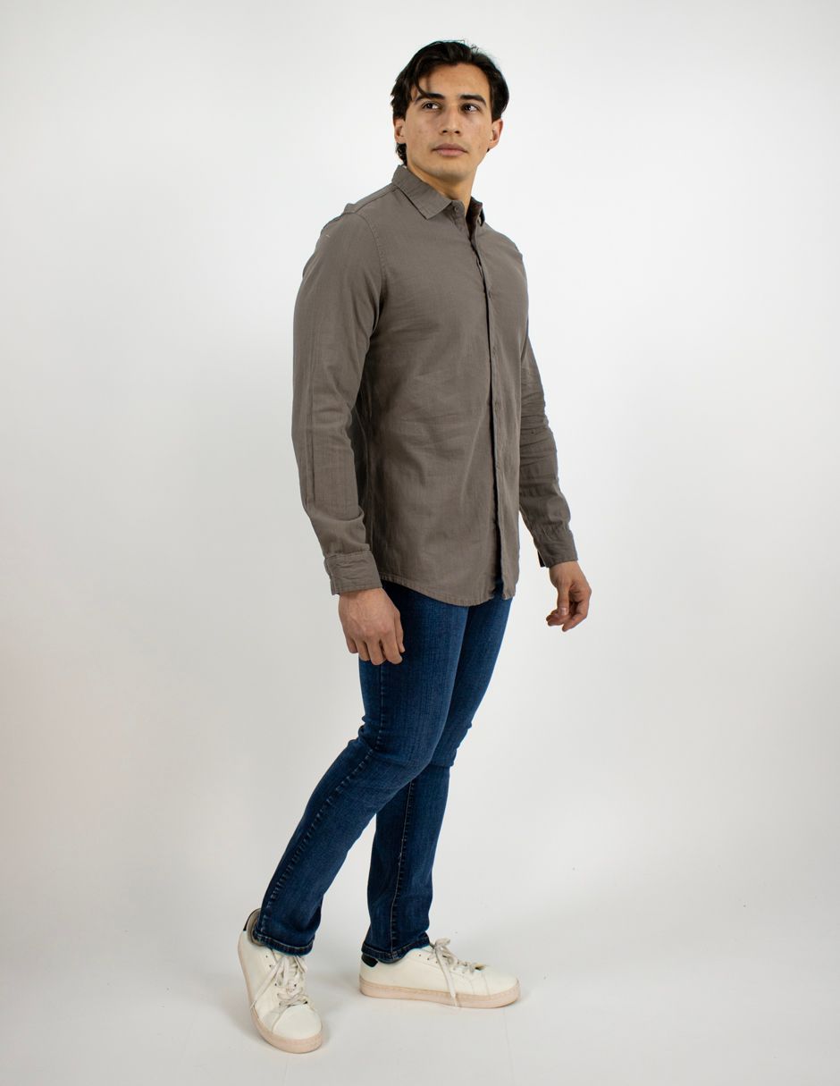 Persona de pie en un estudio, vestida con una camisa gris, vaqueros azules y zapatillas blancas.