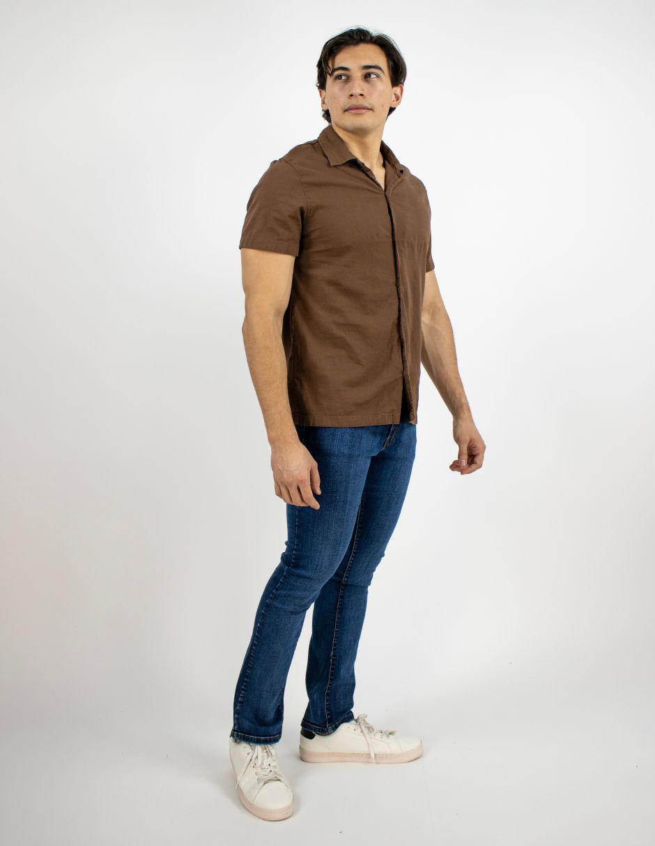 Hombre de pie con camisa marrón, vaqueros azules y zapatillas blancas sobre un fondo blanco liso.