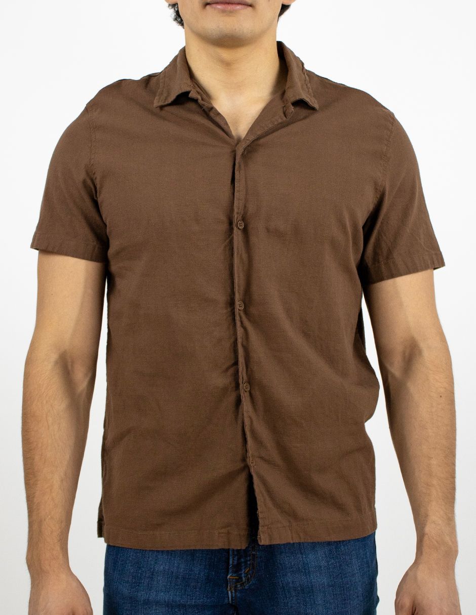 Hombre con camisa marrón de manga corta abotonada y vaqueros azules sobre un fondo blanco.