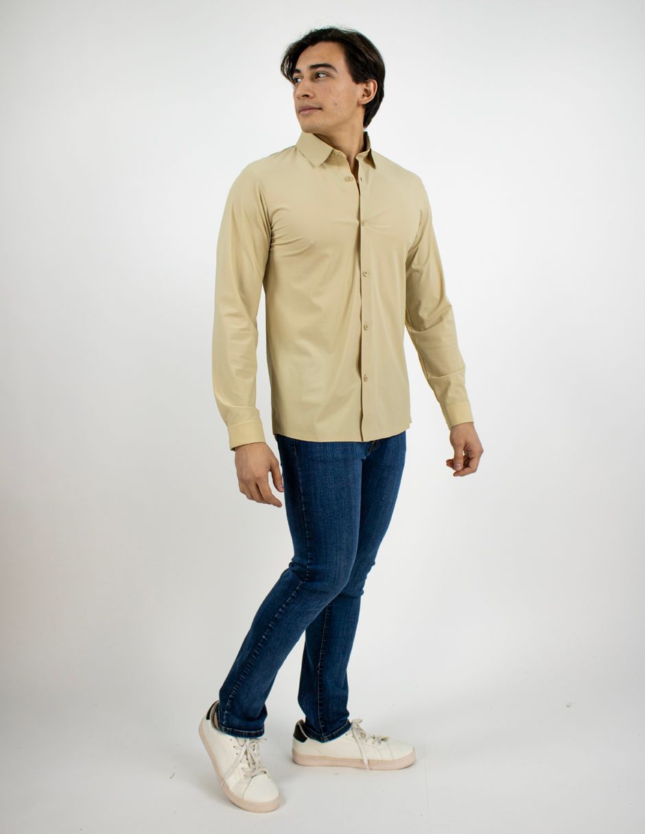 Hombre con camisa beige abotonada, vaqueros azules y zapatillas blancas posando sobre un fondo blanco.