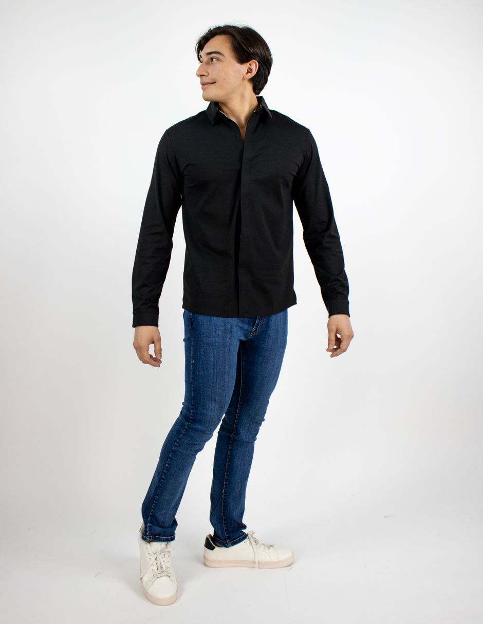 Hombre con camisa negra, vaqueros azules y zapatillas blancas, de pie frente a un fondo blanco de estudio.