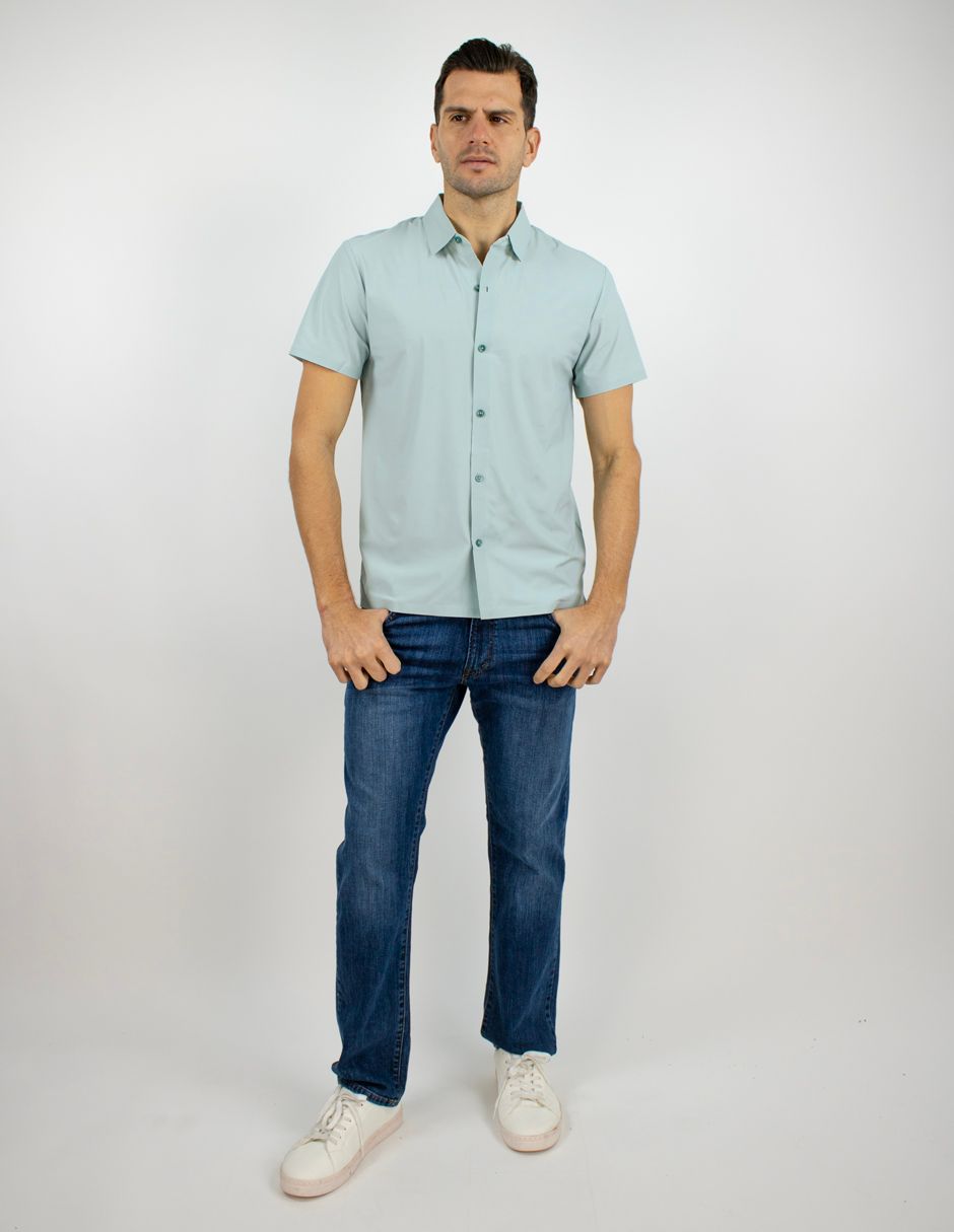 Hombre de pie con una camisa azul claro de manga corta, vaqueros y zapatillas blancas sobre un fondo liso.