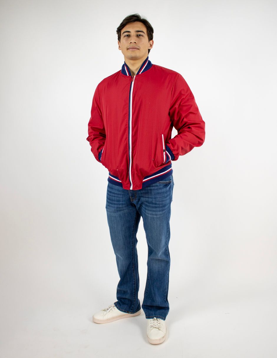 Persona de pie sobre un fondo blanco, vestida con una chaqueta roja, vaqueros azules y zapatillas blancas.