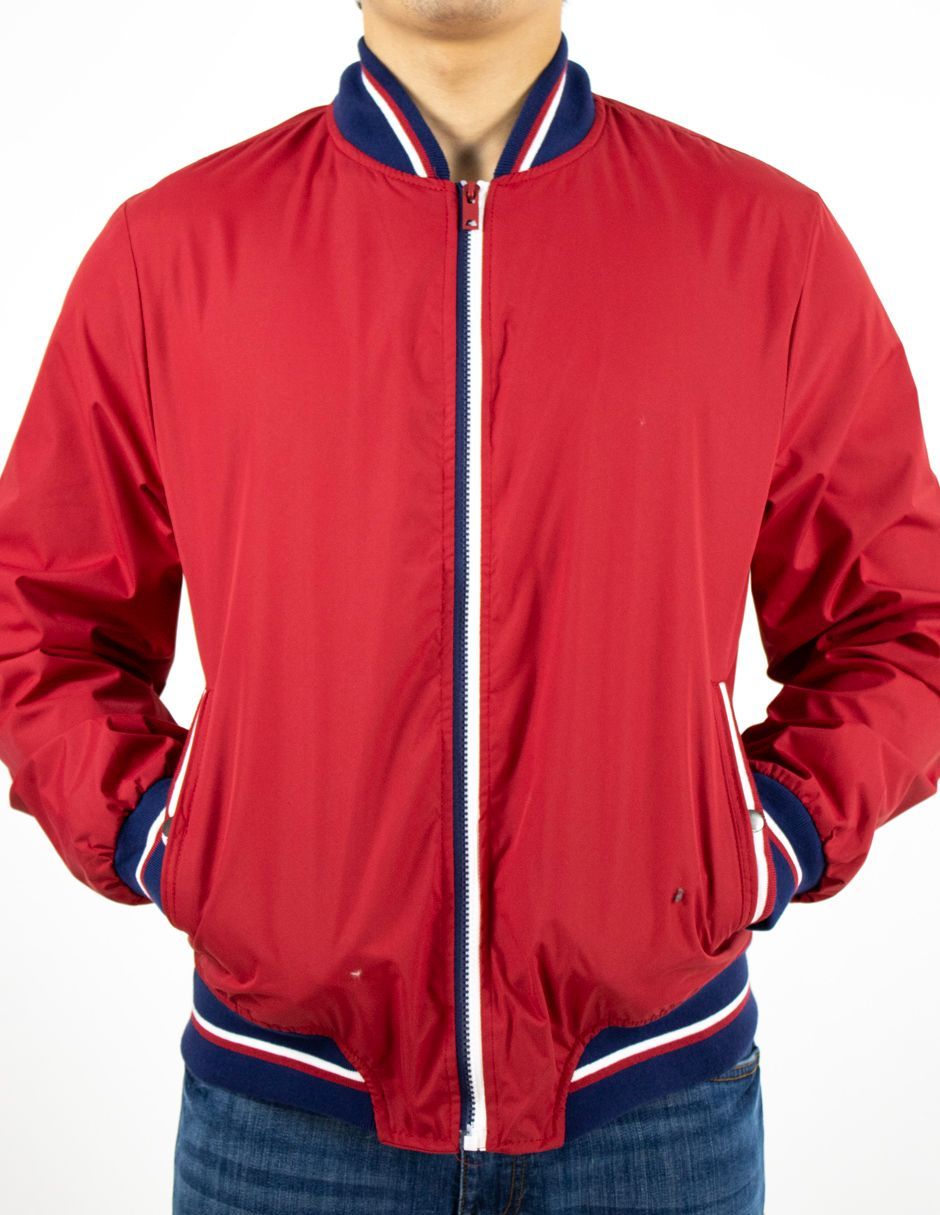 Chaqueta roja con cremallera, cuello, puños y bajo a rayas, usada con las manos en los bolsillos sobre un fondo blanco.