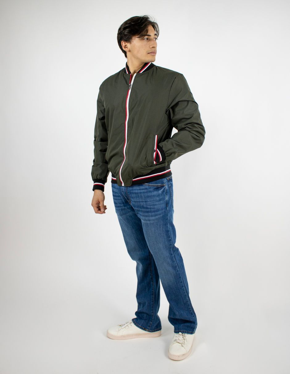 Persona con chaqueta verde oscuro, vaqueros azules y zapatillas blancas de pie sobre un fondo blanco.