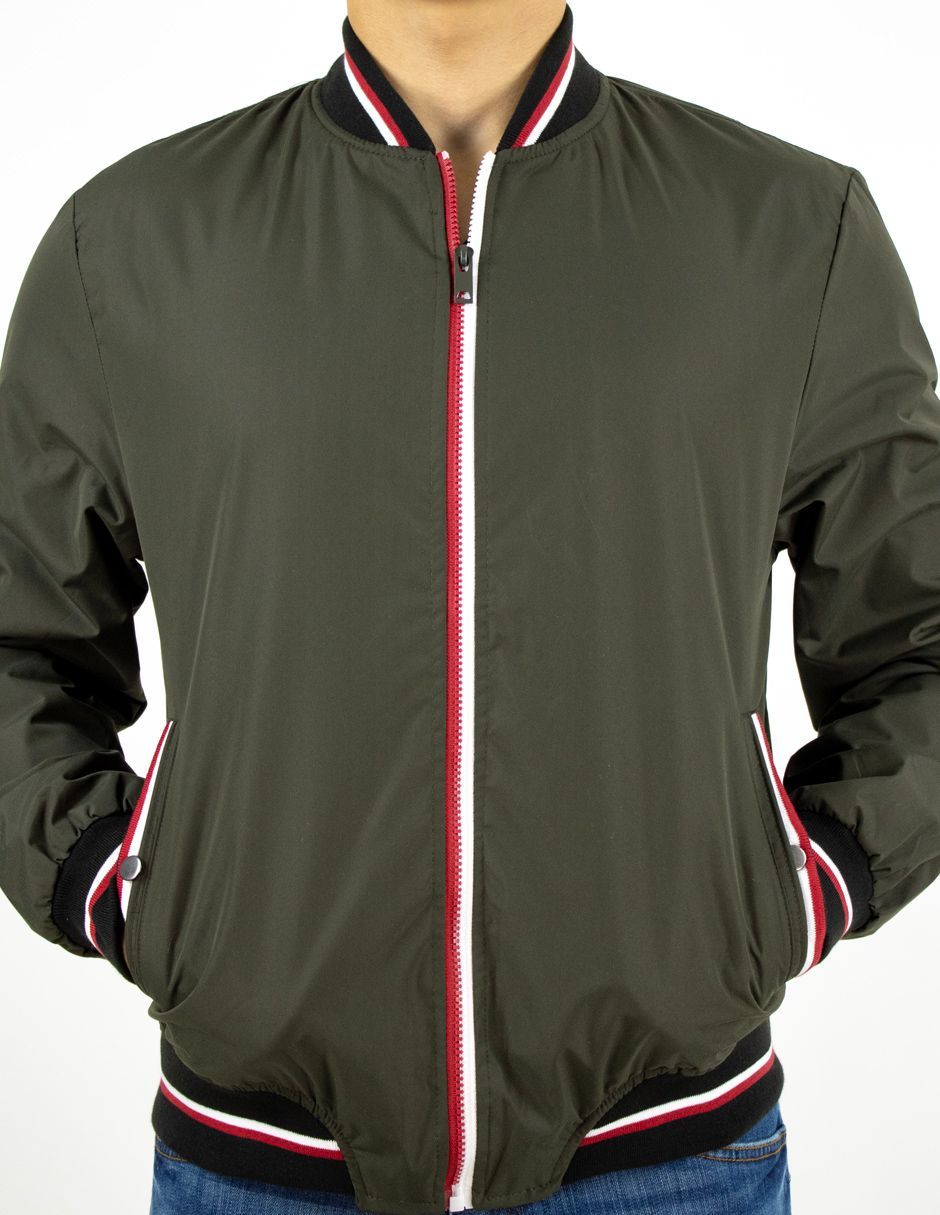 Persona con una chaqueta bomber verde oliva con ribetes de rayas rojas y blancas, con las manos en los bolsillos sobre un fondo blanco.
