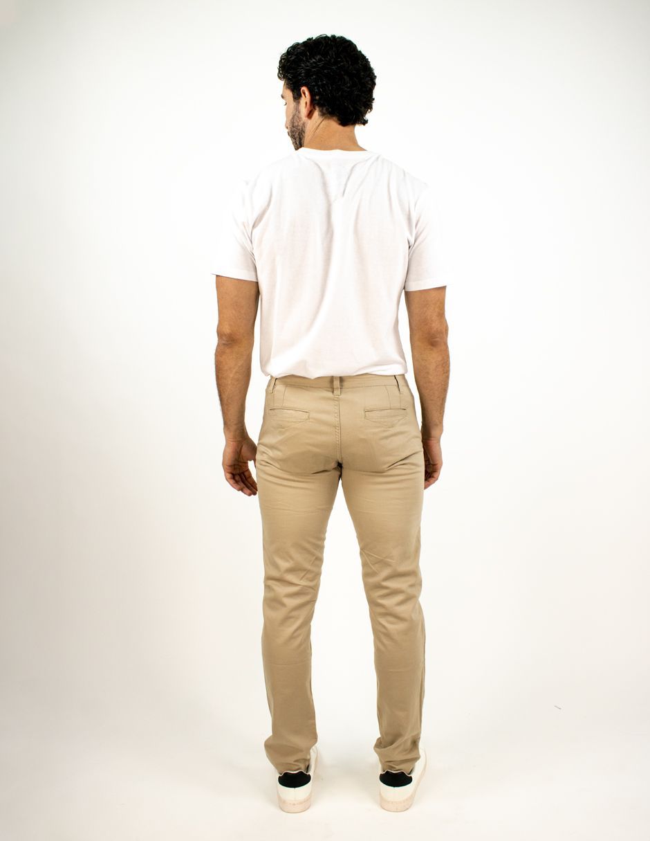 Hombre de pie de espaldas a la cámara, con camiseta blanca, pantalones beige y zapatillas blancas sobre un fondo blanco liso.
