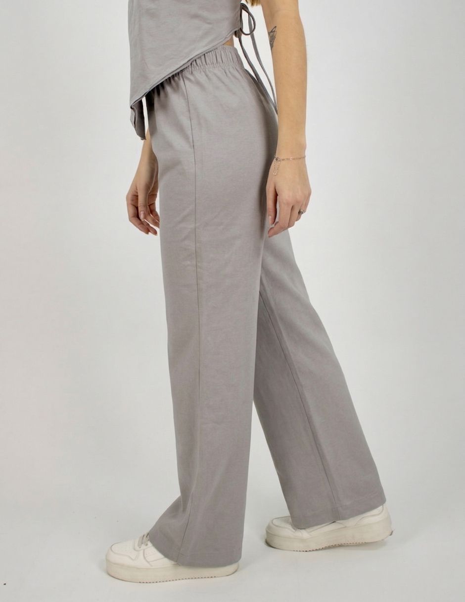 Persona con pantalones anchos de color gris claro y zapatillas blancas de pie sobre un fondo liso.