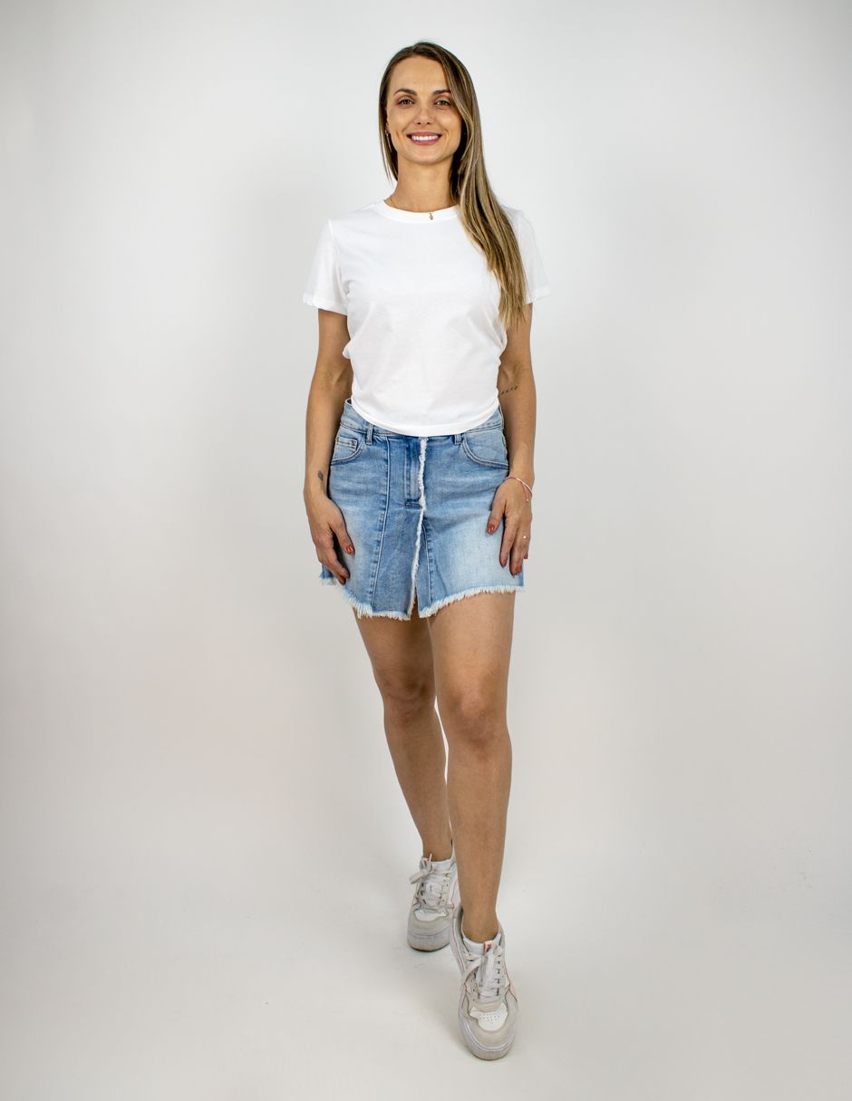 Mujer de pie frente a un fondo blanco, vestida con una camiseta blanca, pantalones cortos vaqueros y zapatillas blancas.
